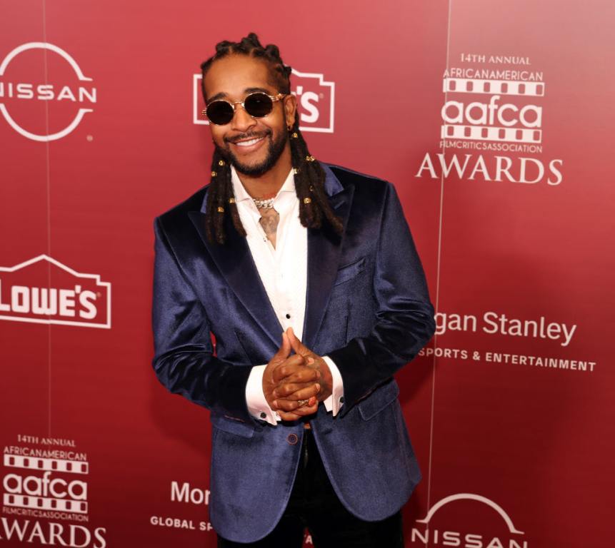 Omarion attends the 14th Annual AAFCA Awards smiling holding his hands wearing a blue blazer and sunglasses.