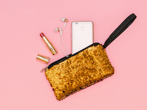 Open lipstick, headphones and phone in a gold-colored bag. Pastel color. Women's accessories. Flat lay.